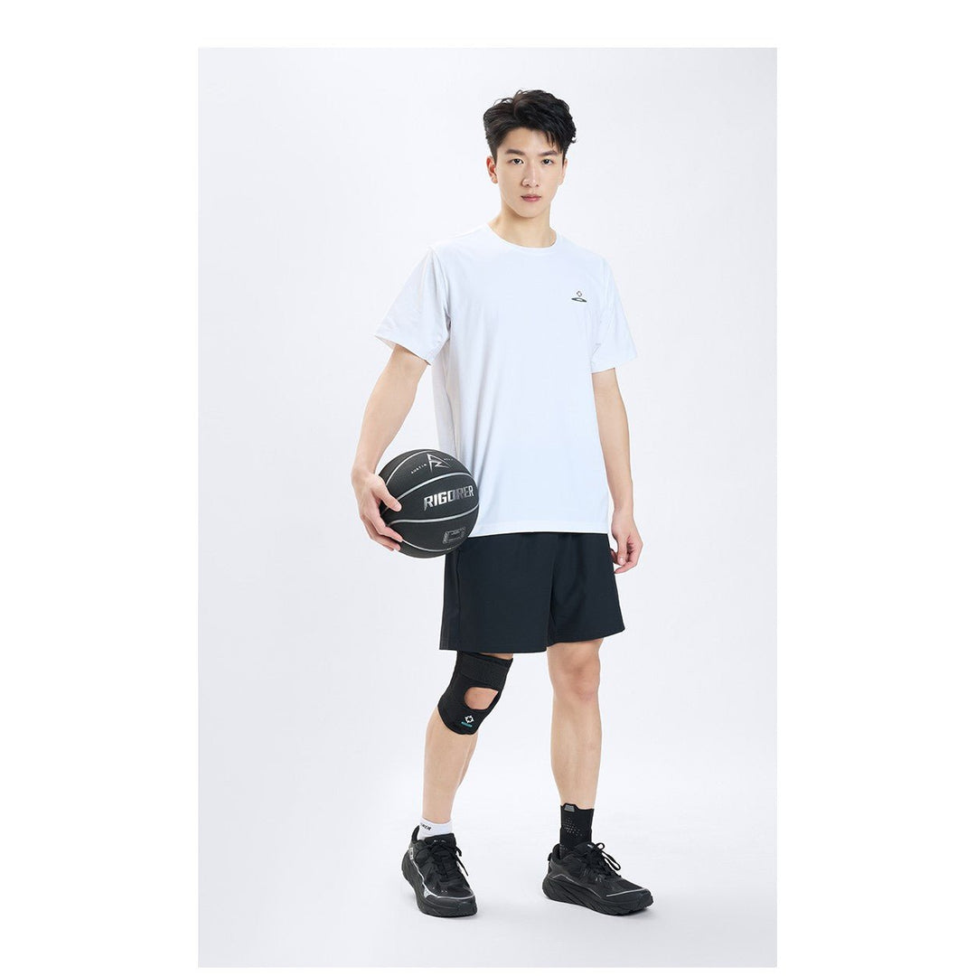 Model in a white tee and black shorts holding a basketball, wearing the Rigorer adult sports knee support brace on his left knee